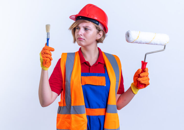 Thinking Young Builder Woman Uniform Holding Roller Brush Looking Paint Brush Her Hand Isolated White Wall